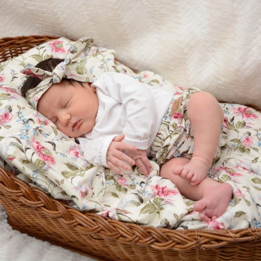 Newborn baby sleeping peacefully in a wicker basket, wrapped in a floral swaddle blanket with matching bow headband.