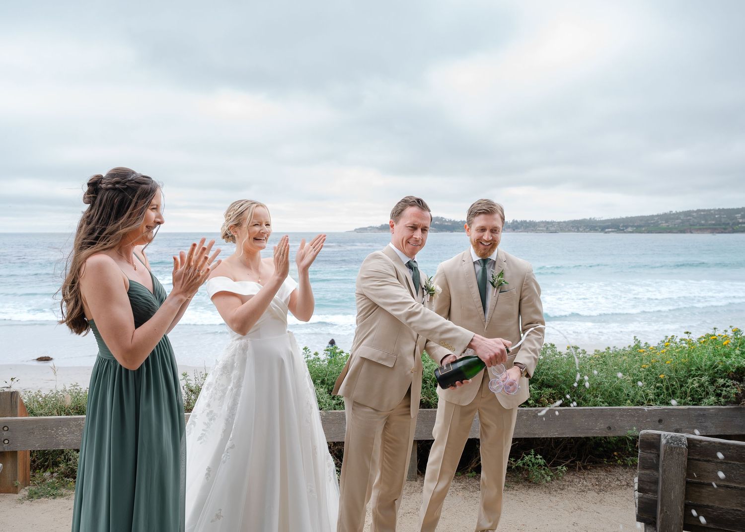 Wedding party celebrates with champagne by the ocean, bride in white gown, groom in tan suit, bridesmaid in sage green dress.