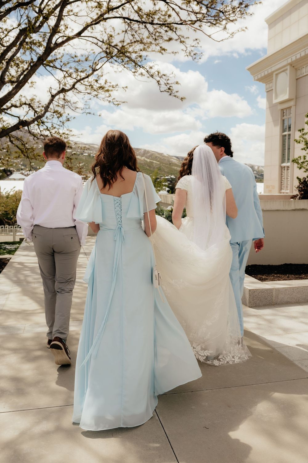 Bride in white gown and bridesmaid in light blue dress walk with groomsmen toward a wedding venue.