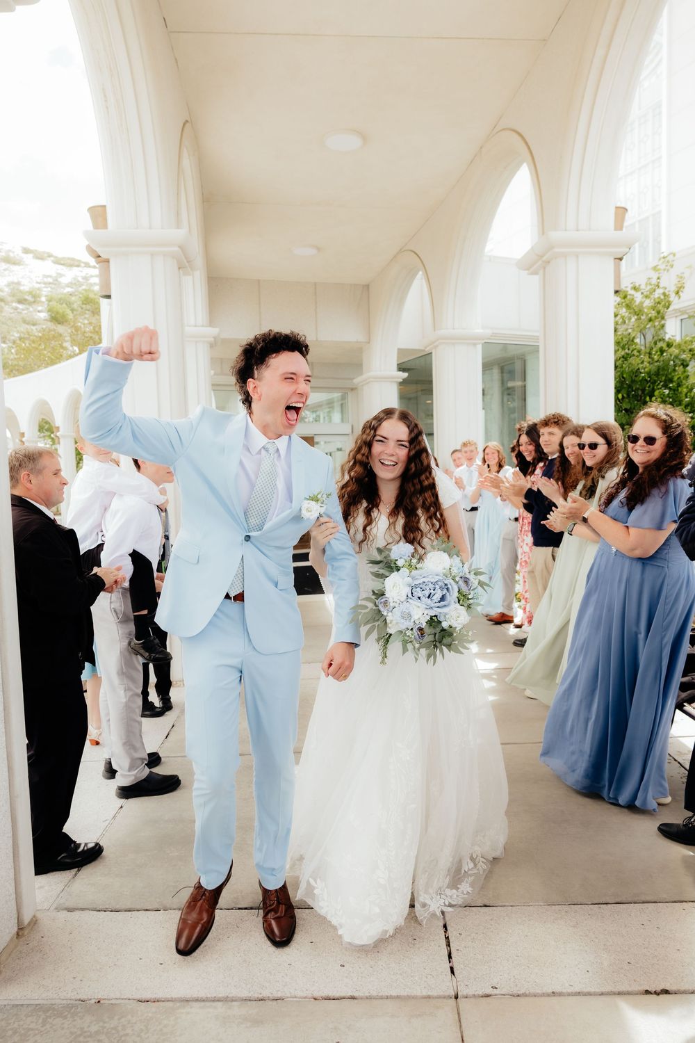 Joyful groom in light blue suit raises fist while bride in white gown holds bouquet as guests celebrate outside white venue.