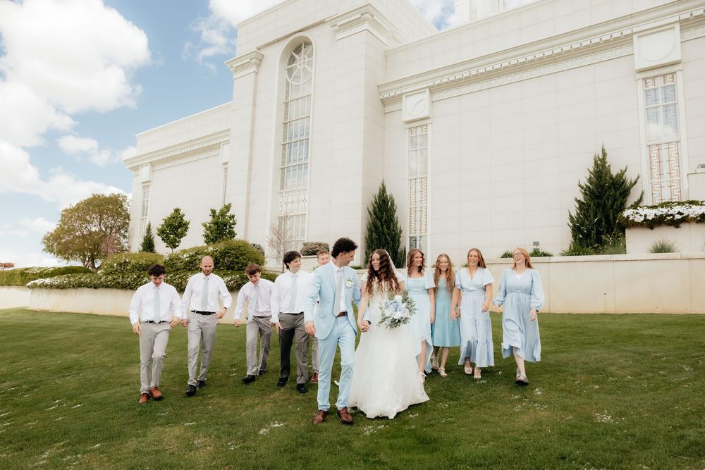 Wedding party walks on green lawn before a grand white temple, groomsmen in gray, bridesmaids in light blue dresses.