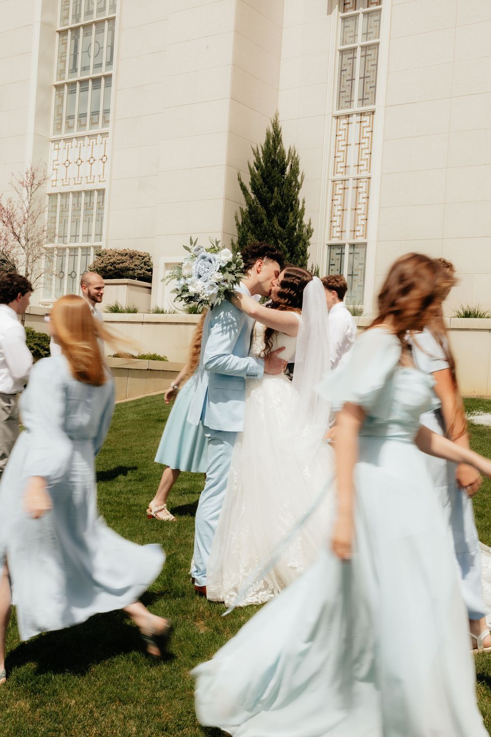 Bride and groom kiss surrounded by bridesmaids in light blue dresses outside a white temple.