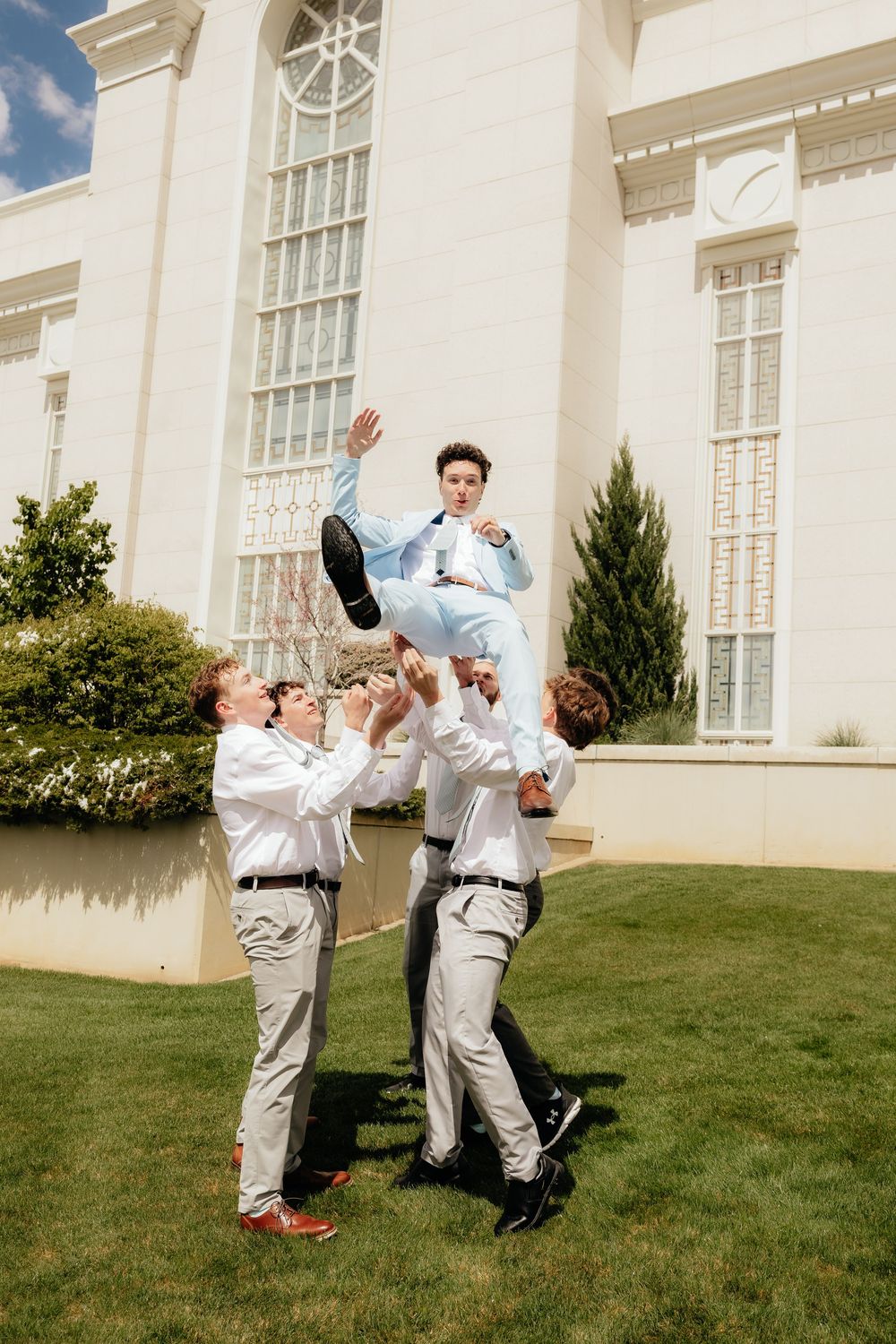 Groomsmen toss groom in light blue suit into the air outside a white wedding venue.