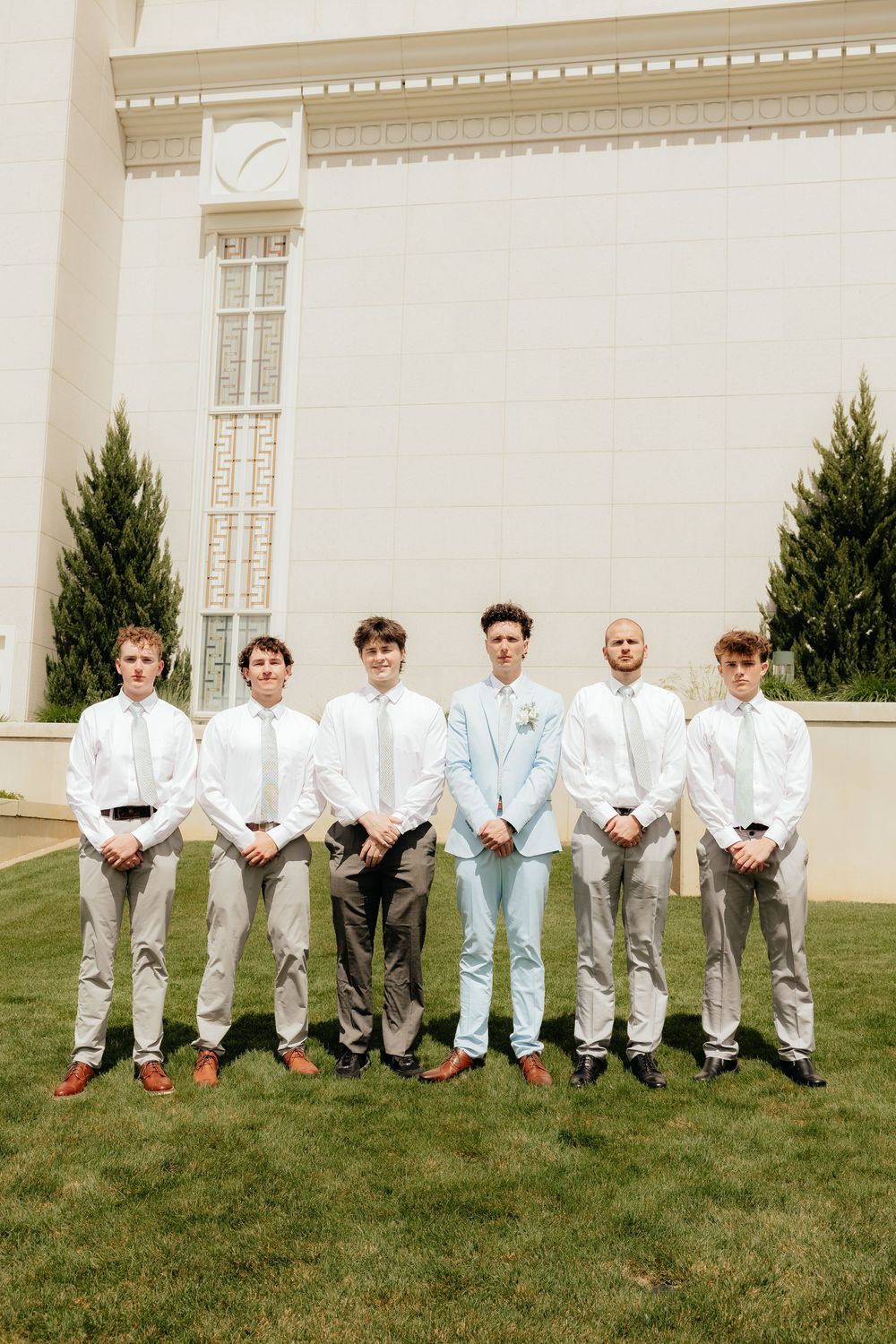Groom in light blue suit stands with six groomsmen in white shirts and ties outside a white building.