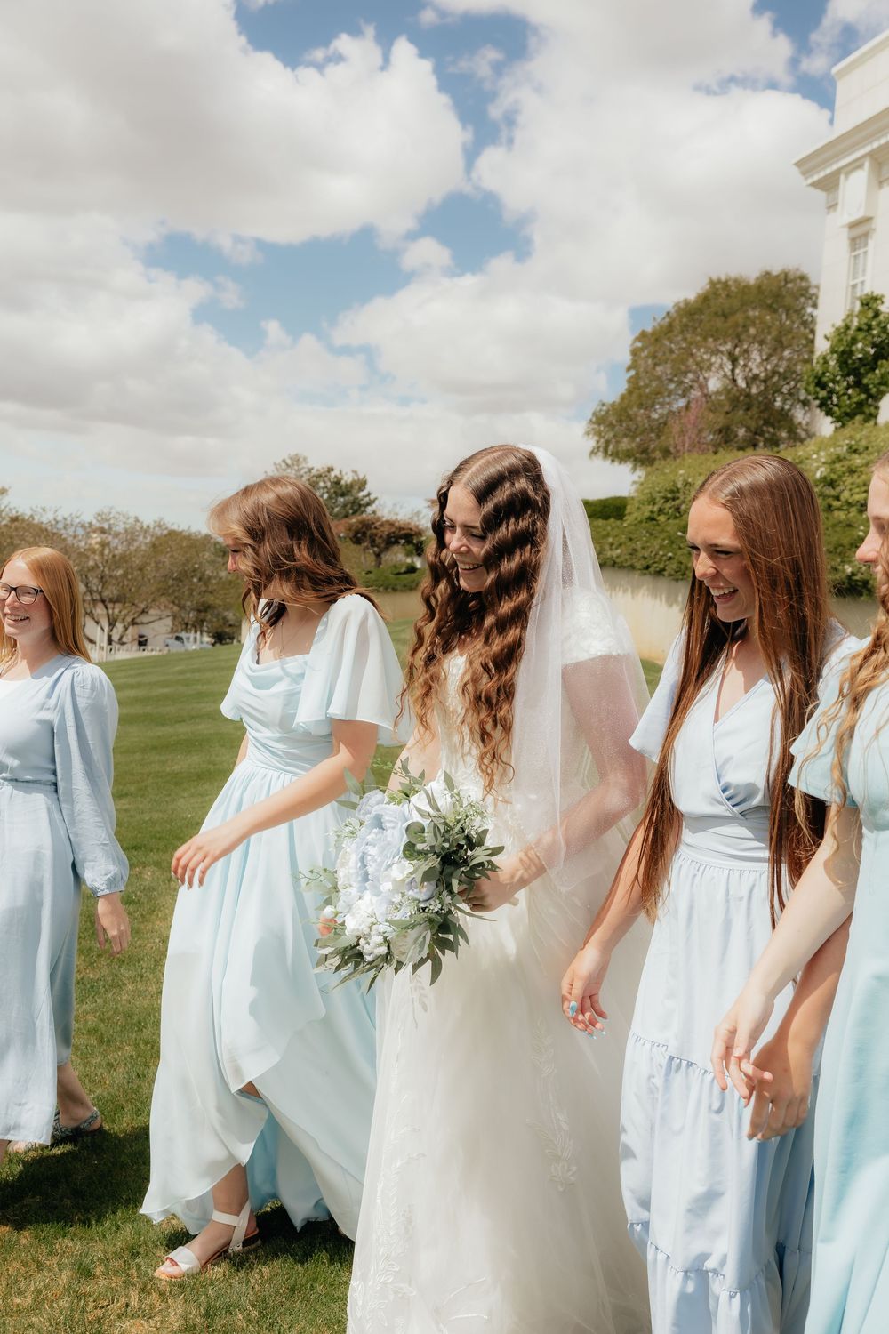 Bride holding white bouquet walks with bridesmaids in light blue dresses on a sunny wedding day.