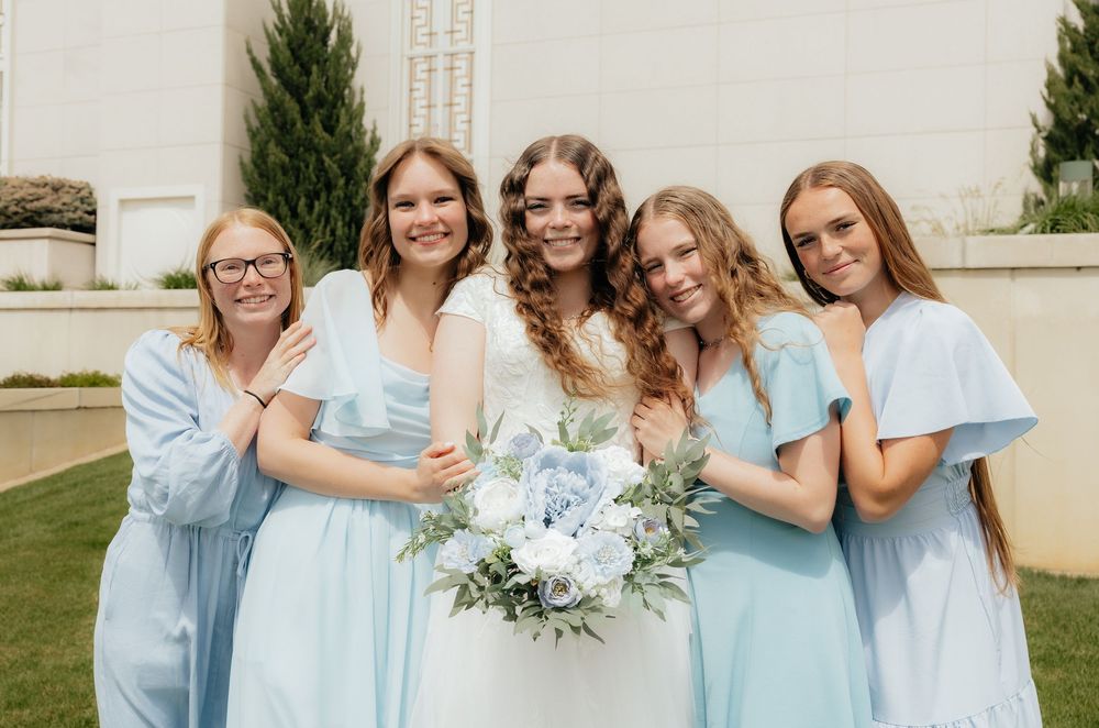 Bride in white gown poses with four bridesmaids in light blue dresses holding a white and blue floral bouquet outdoors.