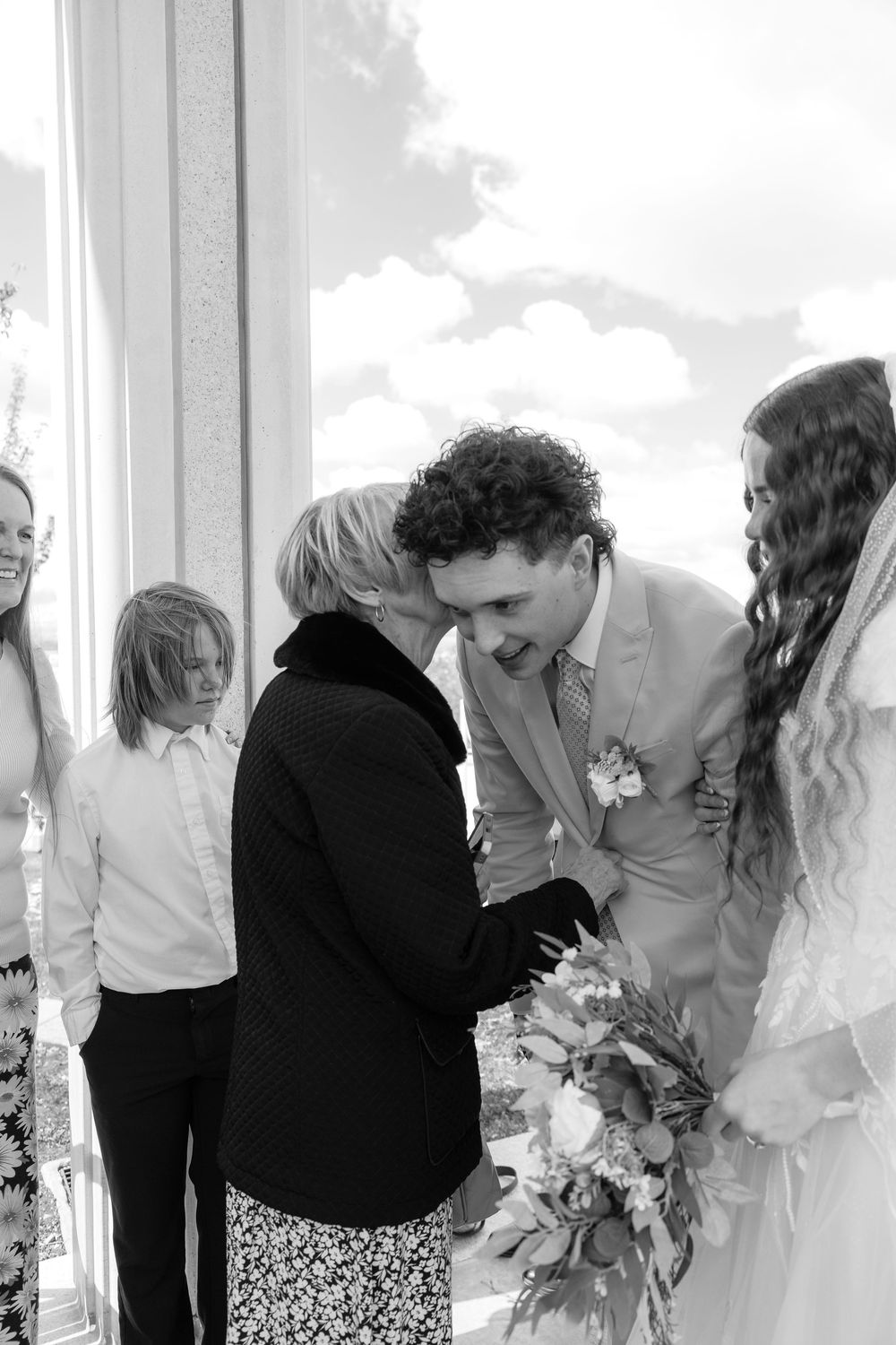 Black and white photo of groom receiving a kiss on the cheek from an elder guest at a wedding ceremony.