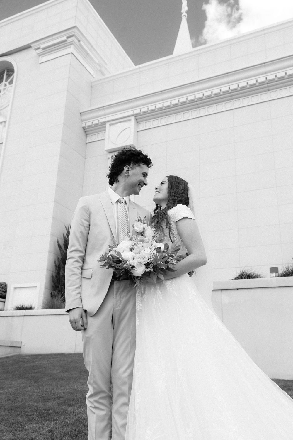 Black and white photo of smiling bride and groom embracing outside a white temple building.