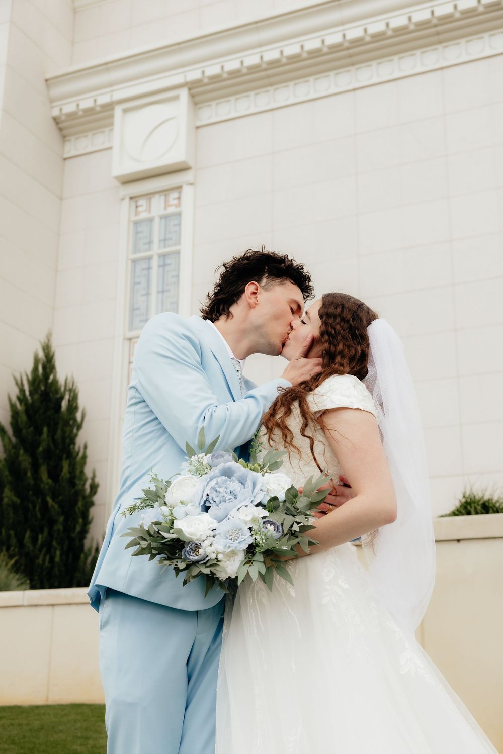 Newlyweds share a romantic kiss outside a white wedding venue, groom in light blue suit holding bridal bouquet.