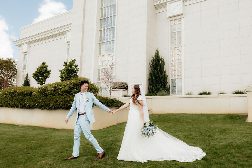 Bride and groom hold hands walking on green lawn outside a white LDS temple on wedding day.
