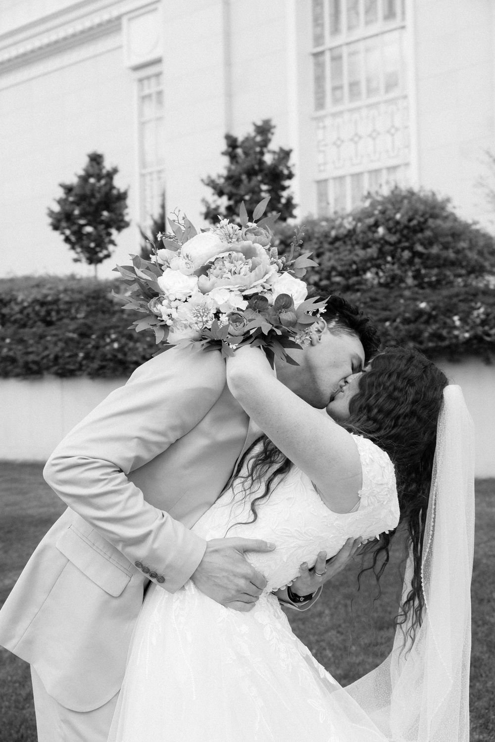 Black and white photo of bride and groom sharing a romantic kiss while holding a floral bouquet outdoors.