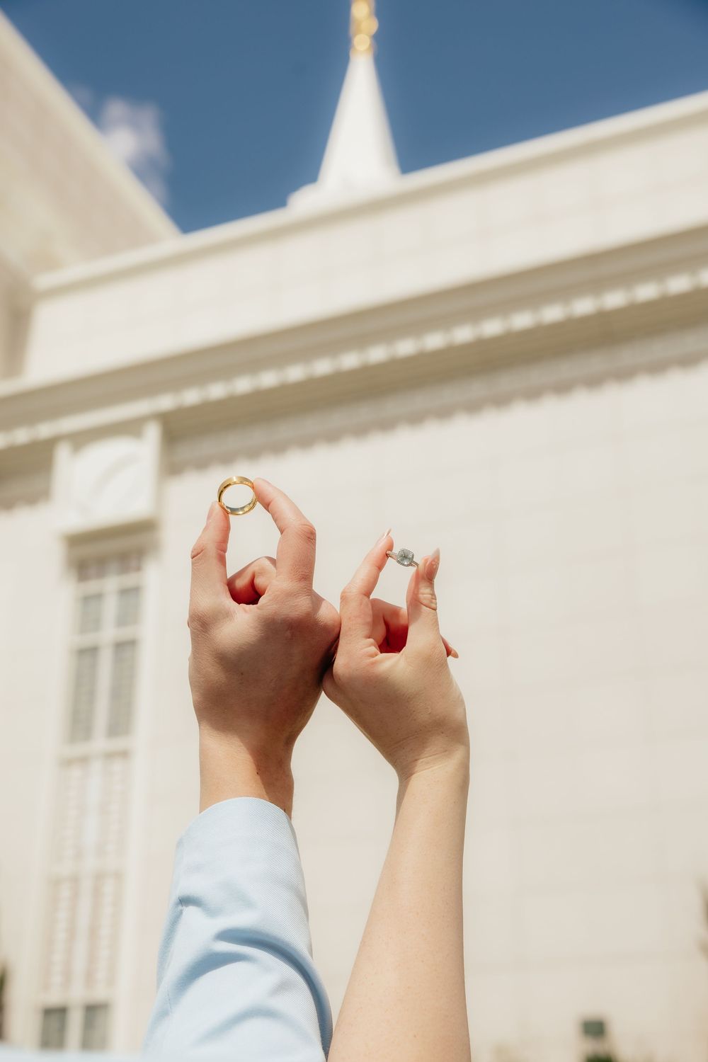 Bride and groom hold up wedding rings in front of a white temple spire against a blue sky.