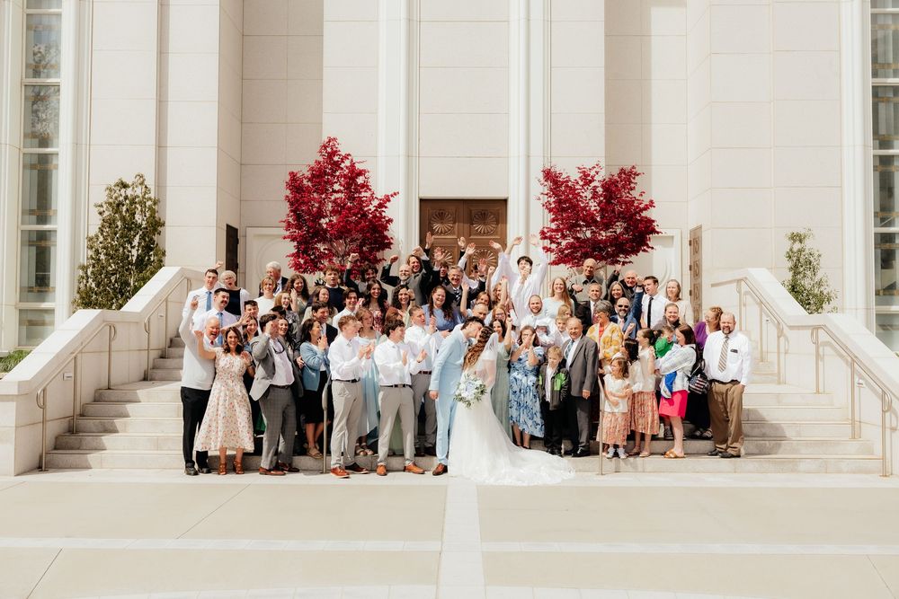 Large wedding group photo on temple steps with red trees, bride and groom surrounded by smiling guests in summer attire.