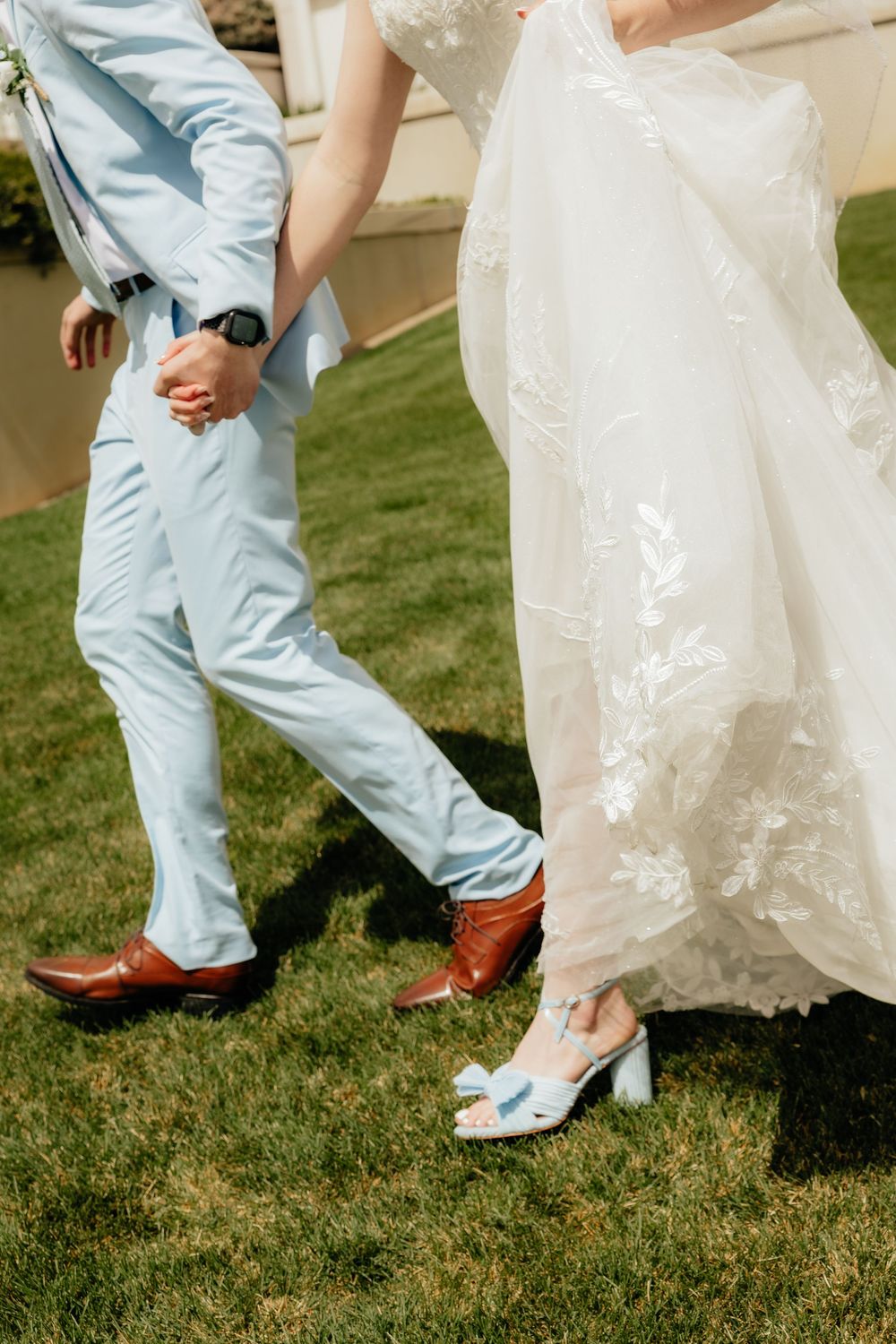 Close-up of bride in white lace gown and groom in light blue suit holding hands on grass.