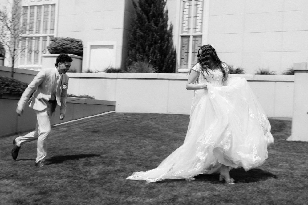 Black and white photo of a bride in a flowing gown and groom in a suit playfully running outdoors.