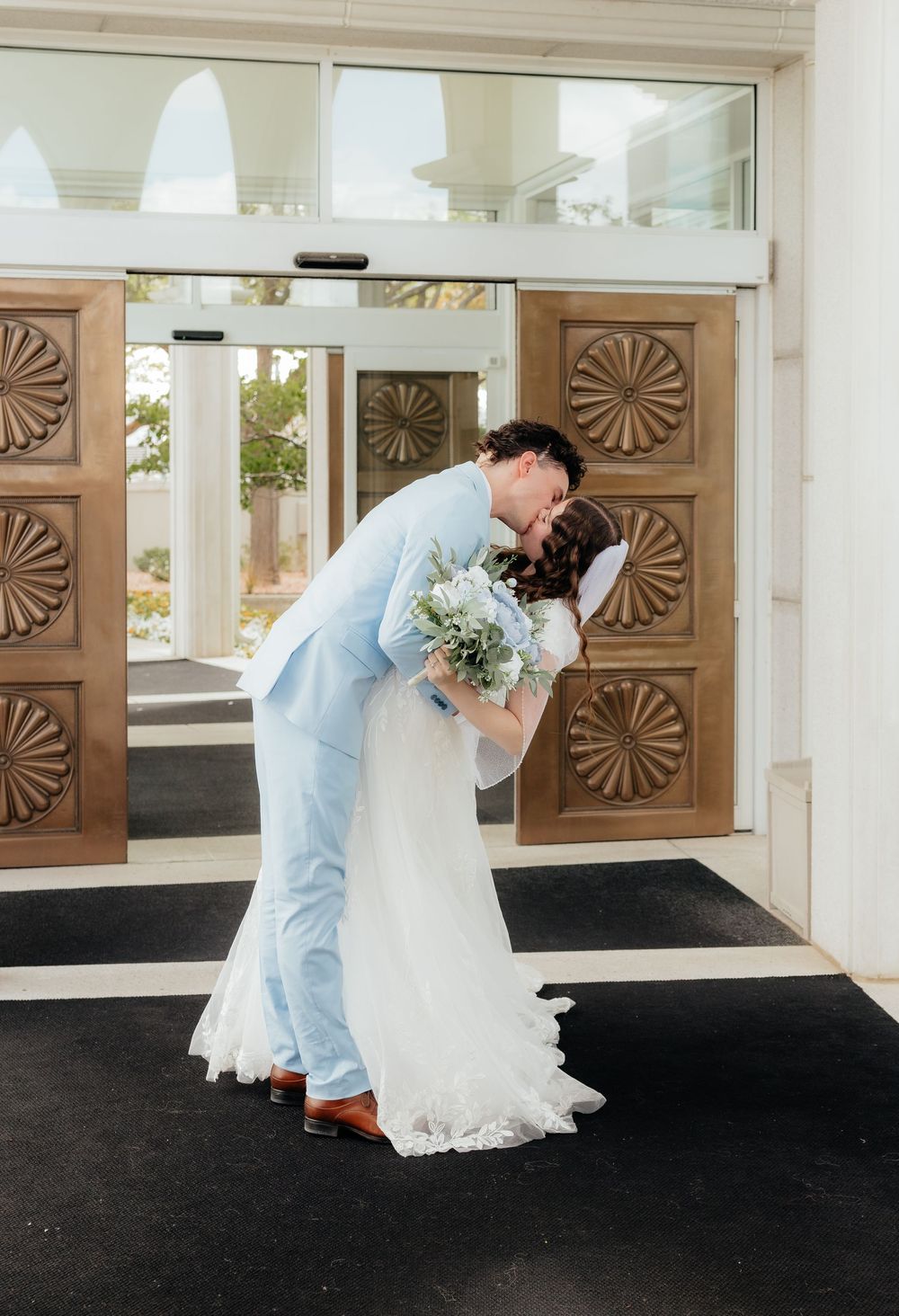 Bride and groom share a romantic dip kiss outside ornate wooden doors in wedding attire.