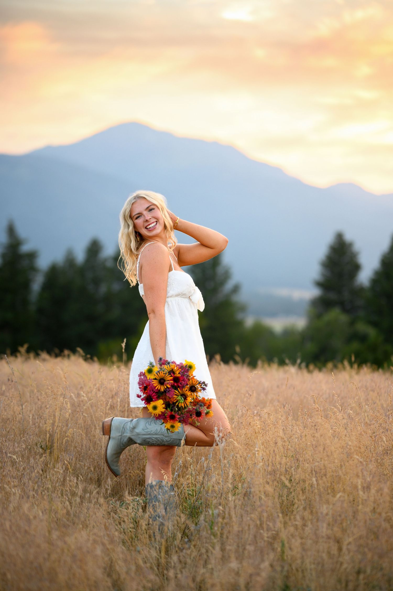 Joyful figure in white dress holds colorful wildflower bouquet while standing in golden field at sunset with mountains behind.