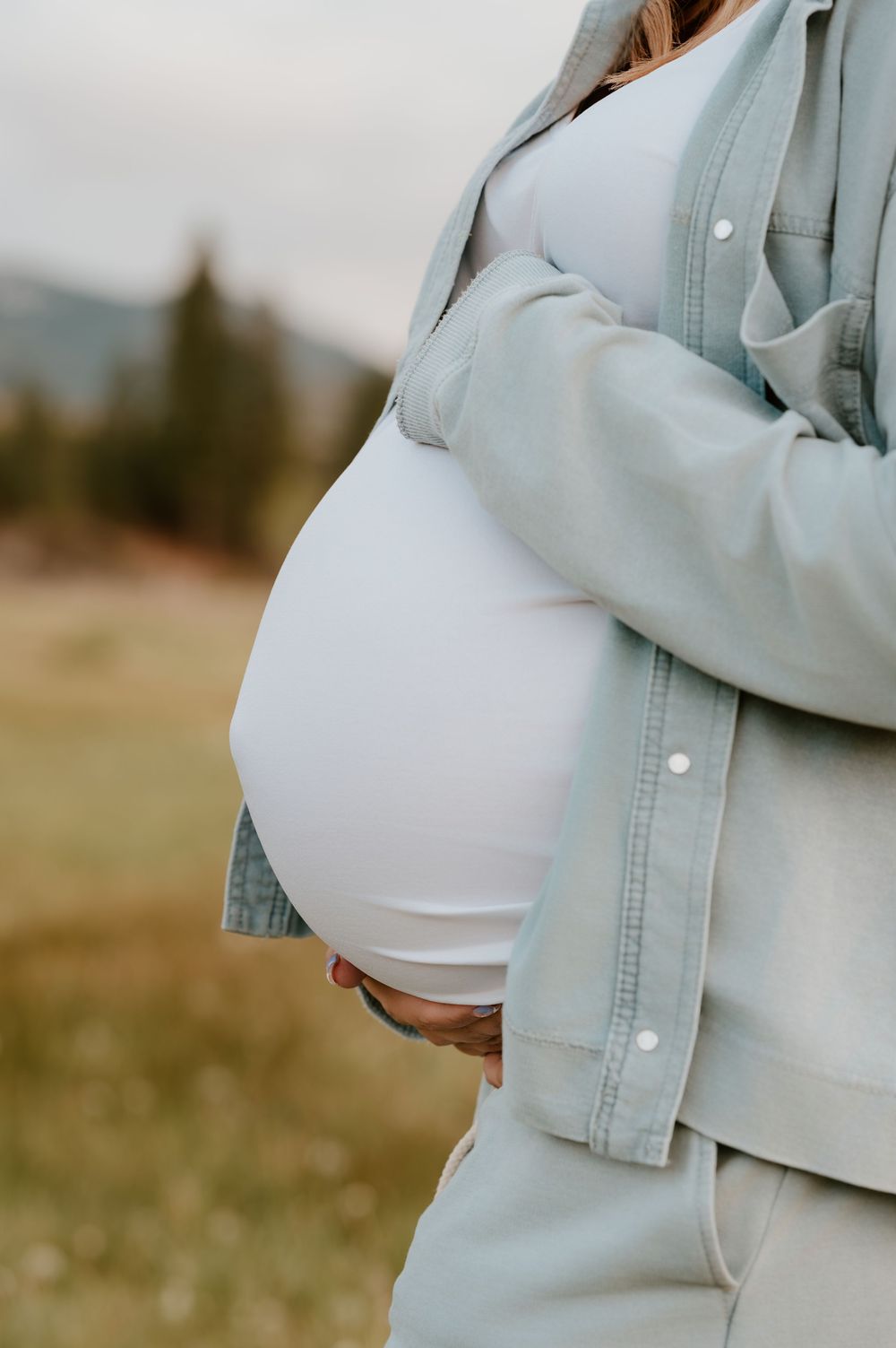 Pregnant woman in light blue denim shirt cradling baby bump outdoors in a green field.