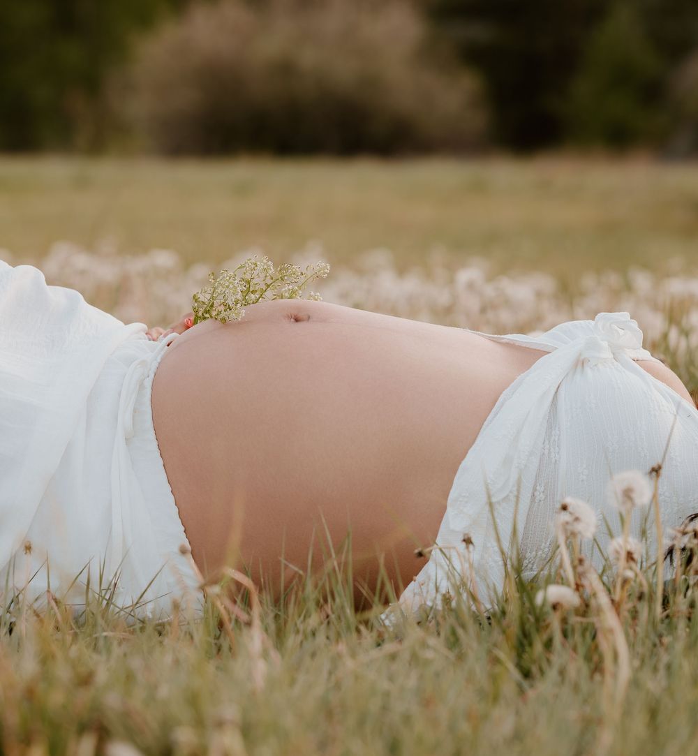 Pregnant woman in white dress lying in a wildflower meadow, showcasing her bare baby bump outdoors.
