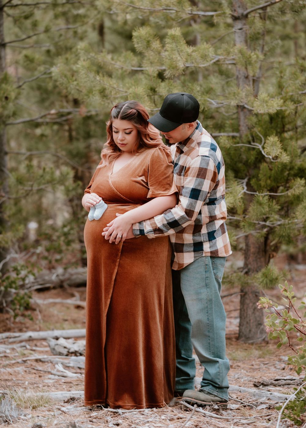 Couple in forest during maternity shoot, man in plaid shirt holds baby shoe near pregnant womans belly in rust dress.