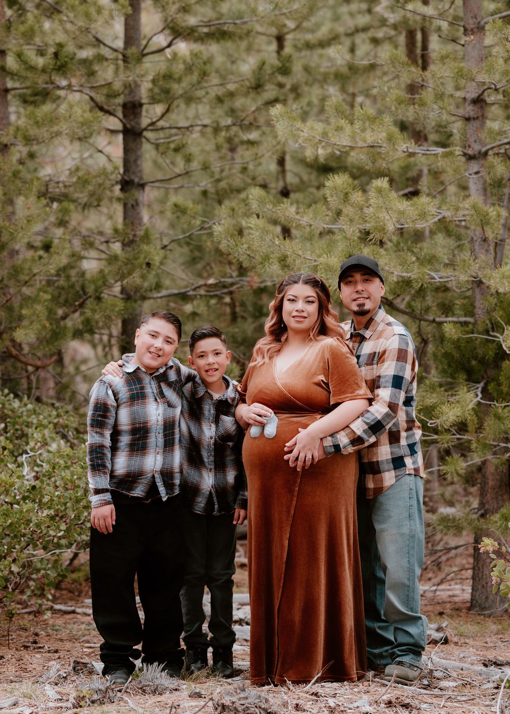 Family maternity photo session in pine forest with pregnant mother in rust velvet dress, father, and two boys in plaid shirts.