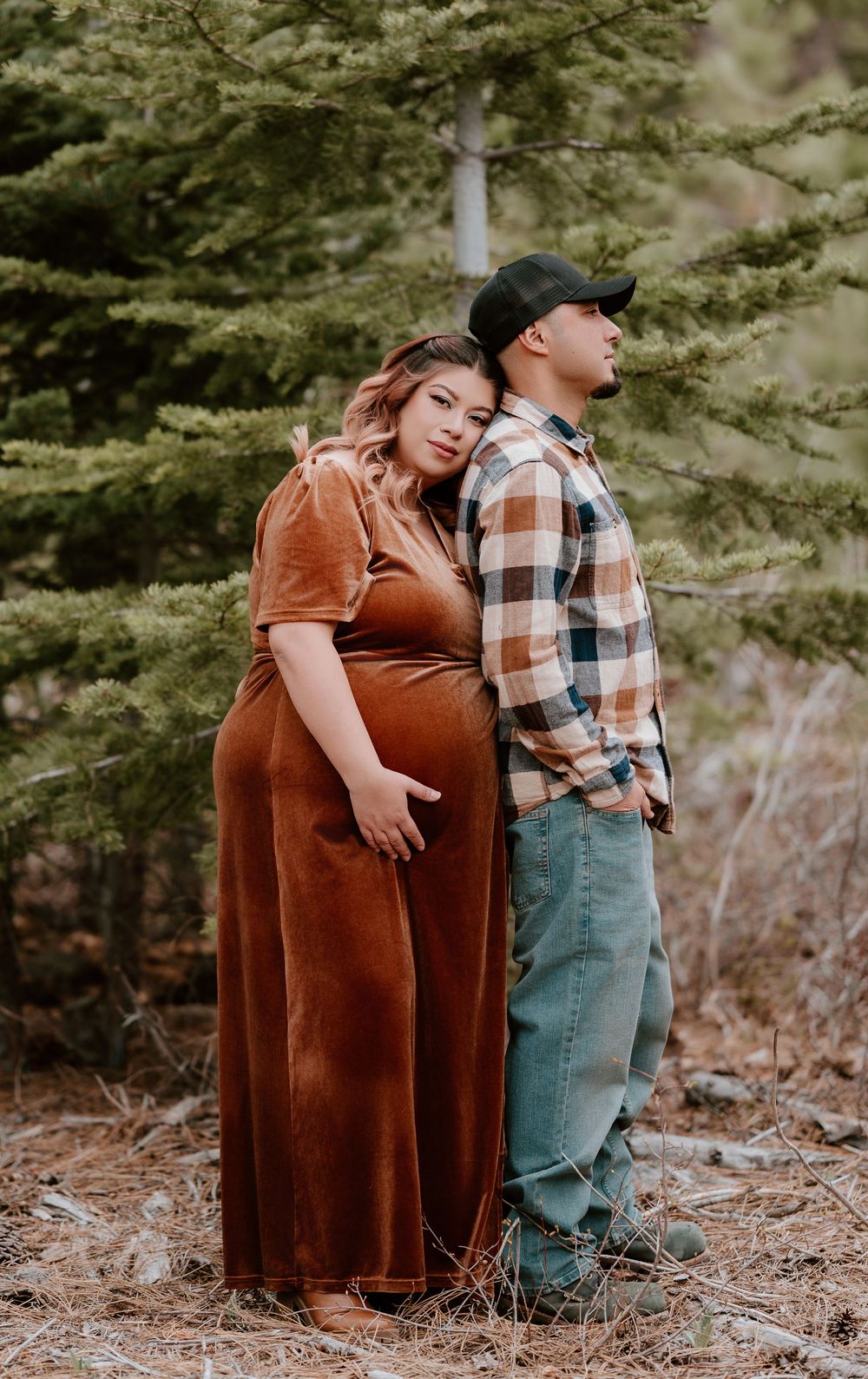 Pregnant woman in rust velvet dress leans on partner in plaid shirt during outdoor maternity photo session in pine forest.