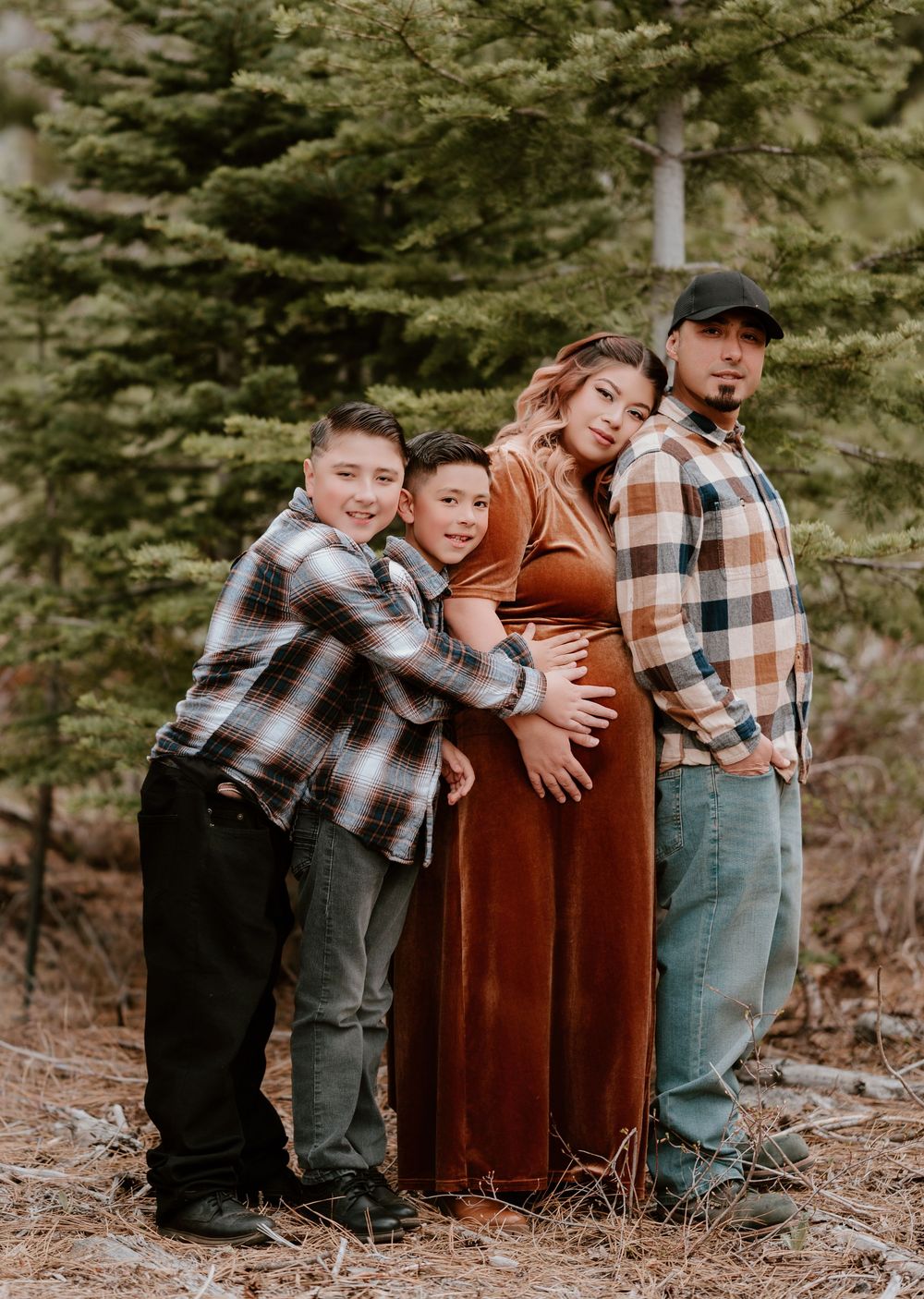 Family maternity photo with two boys, pregnant mother in rust dress, and father among pine trees.