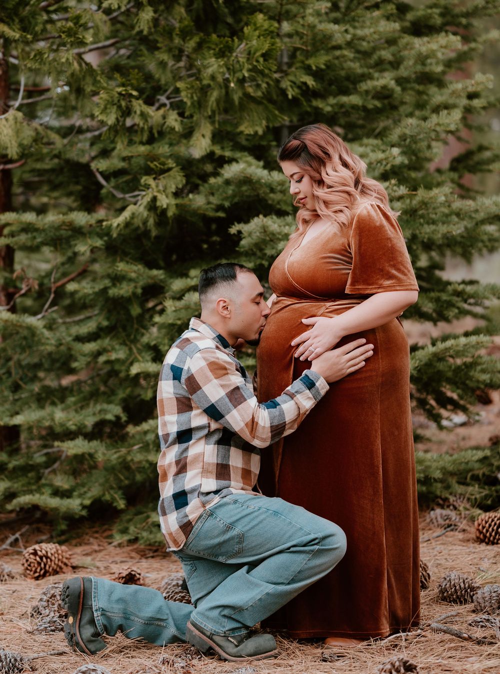 Expectant father kneeling and kissing pregnant womans baby bump during outdoor maternity photo session among pine trees.