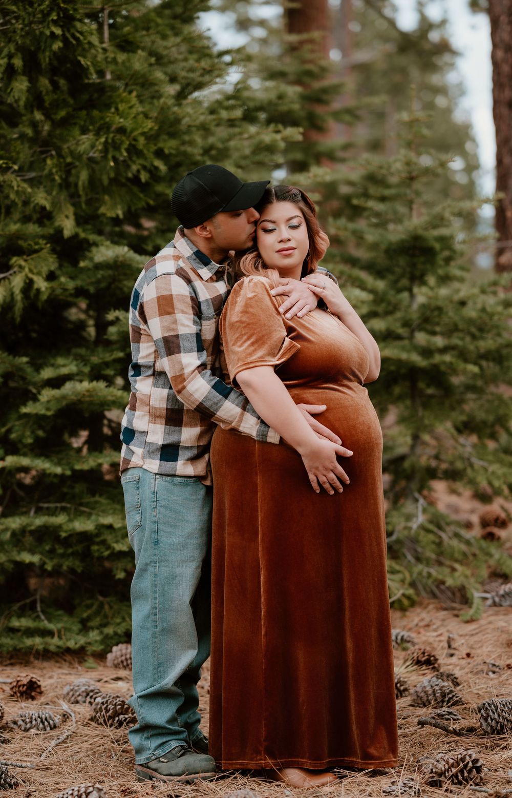Couple maternity photo in forest, woman in rust velvet gown, man in flannel kissing her cheek among pine trees.