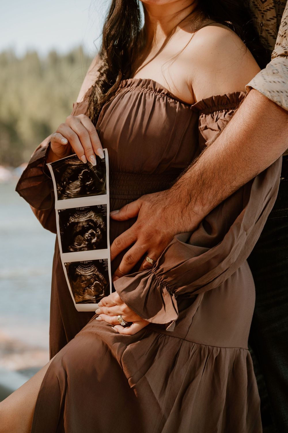Pregnant couple holding ultrasound photos outdoors near water, woman in brown off-shoulder dress, maternity announcement photo.