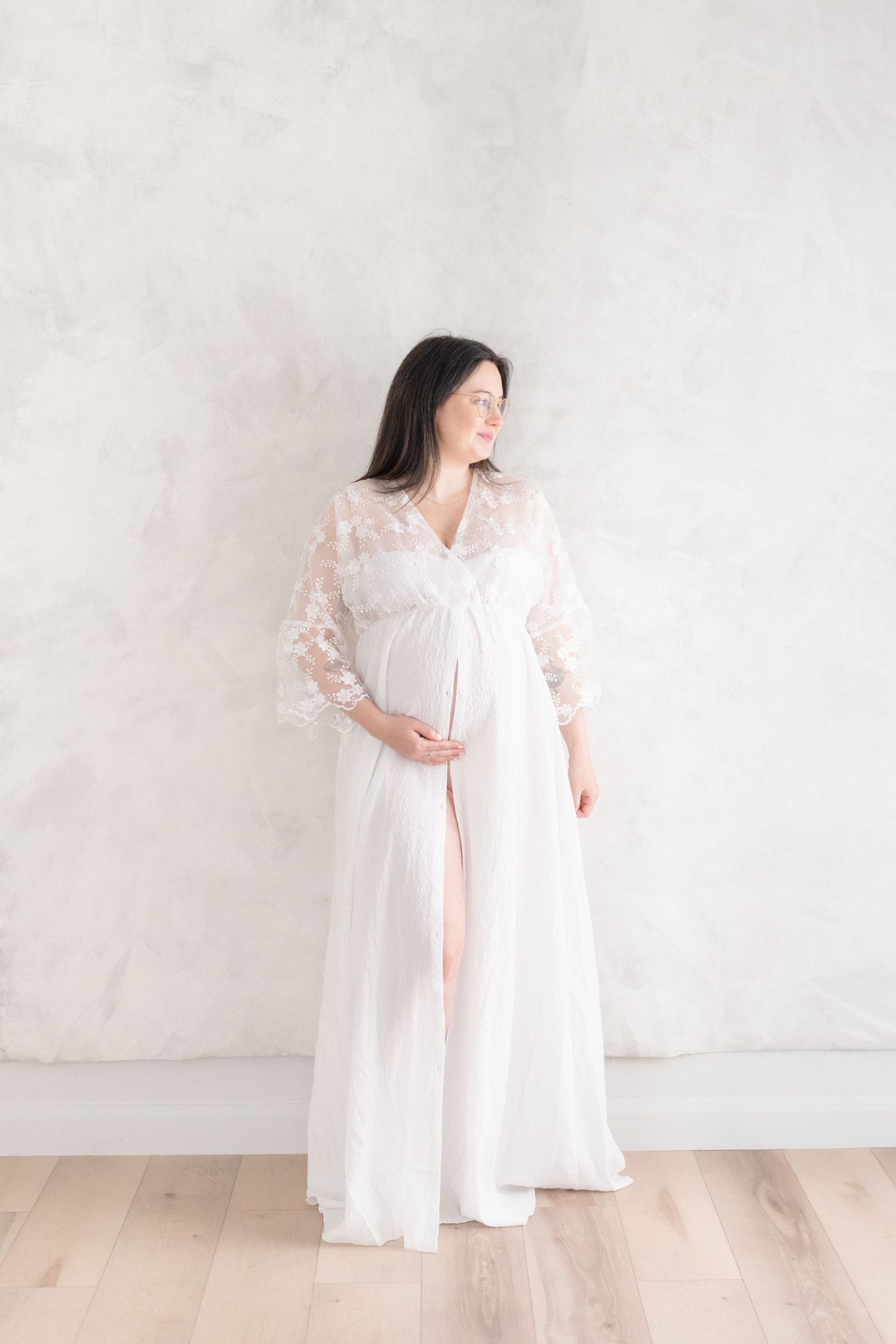 Portrait artistique d'une future maman dans une fabuleuse robe fluide de dentelle et de tulle blanc dans la lumière naturelle du studio