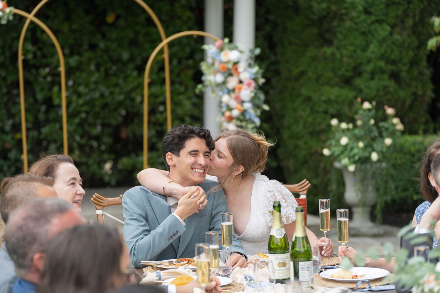 Couple sharing a romantic moment at an outdoor wedding reception dinner table with floral arch backdrop.