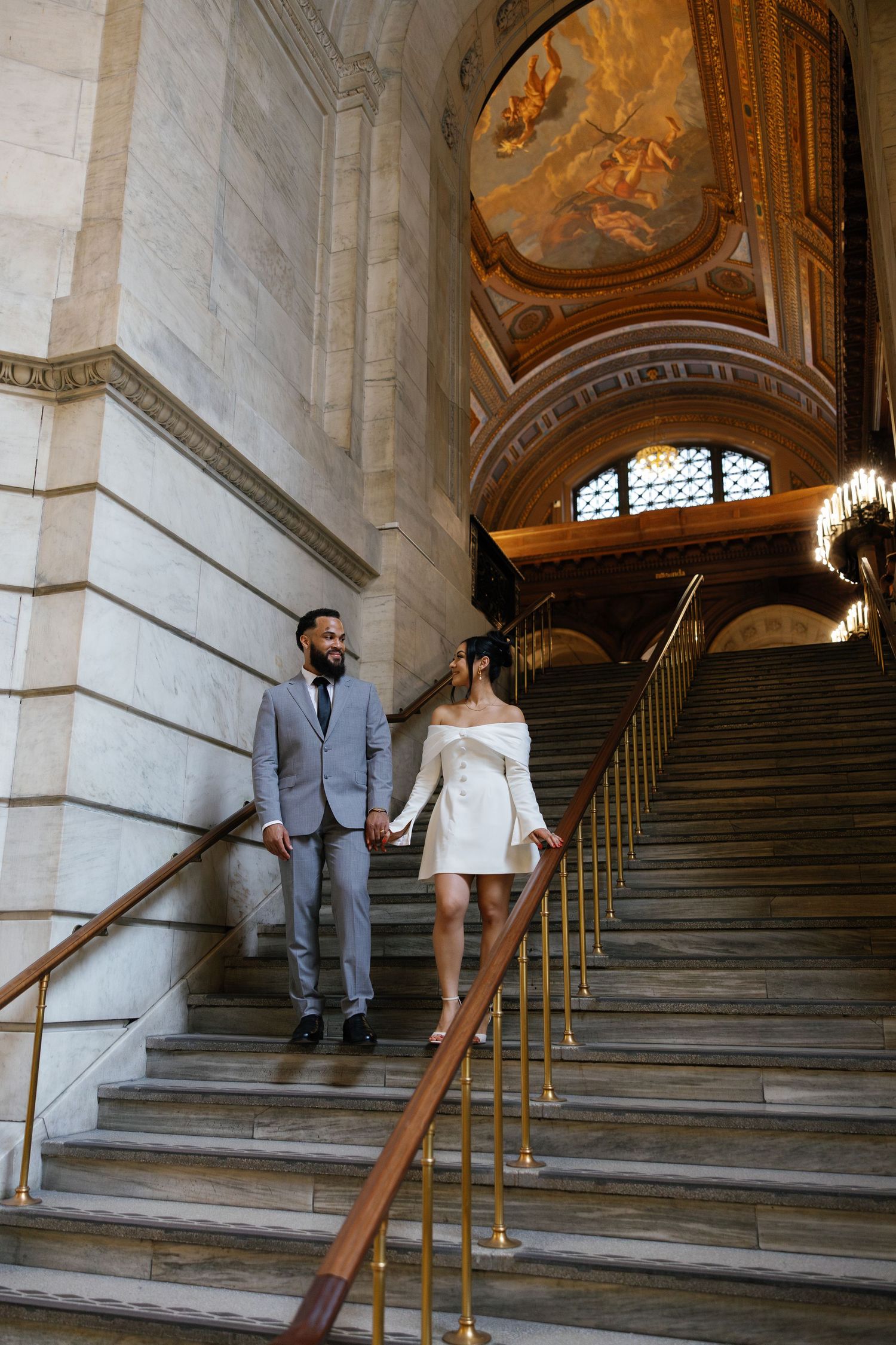 Elegant couple holding hands descending grand marble staircase in ornate building with golden ceiling mural.