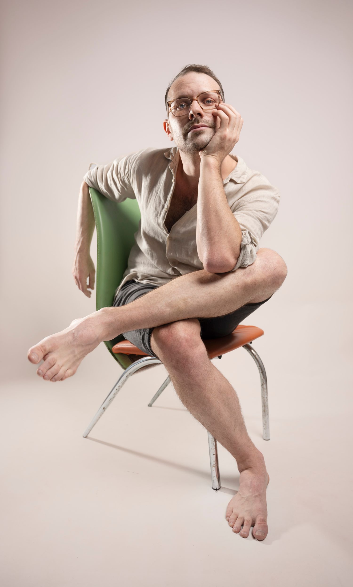 Barefoot man in glasses sitting cross-legged on a green and orange chair against a light background.