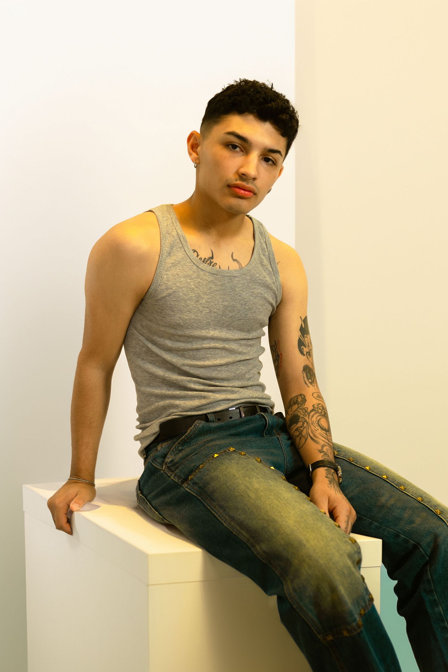 Young man with tattoos sits on white cube wearing gray tank top and jeans in a studio setting.