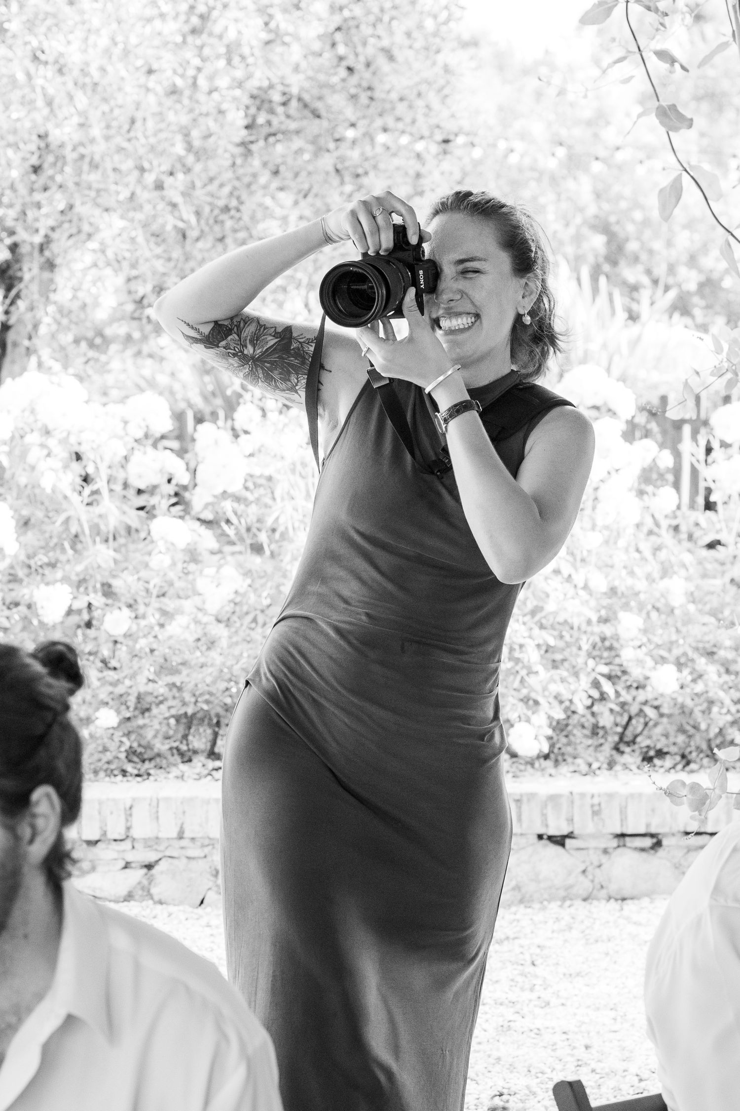 A smiling woman in a sleeveless dress photographs an outdoor wedding event with a DSLR camera.