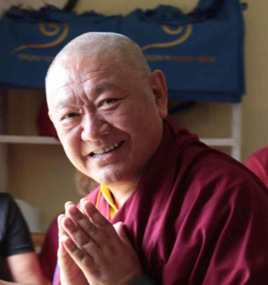 Smiling Buddhist monk in maroon robes with hands in prayer gesture, bald head, seated indoors.