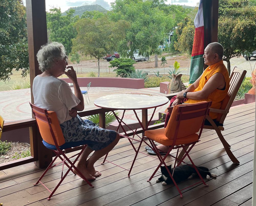 A Buddhist monk in orange robes sits with a person on a wooden deck, sharing a peaceful conversation at a small table.