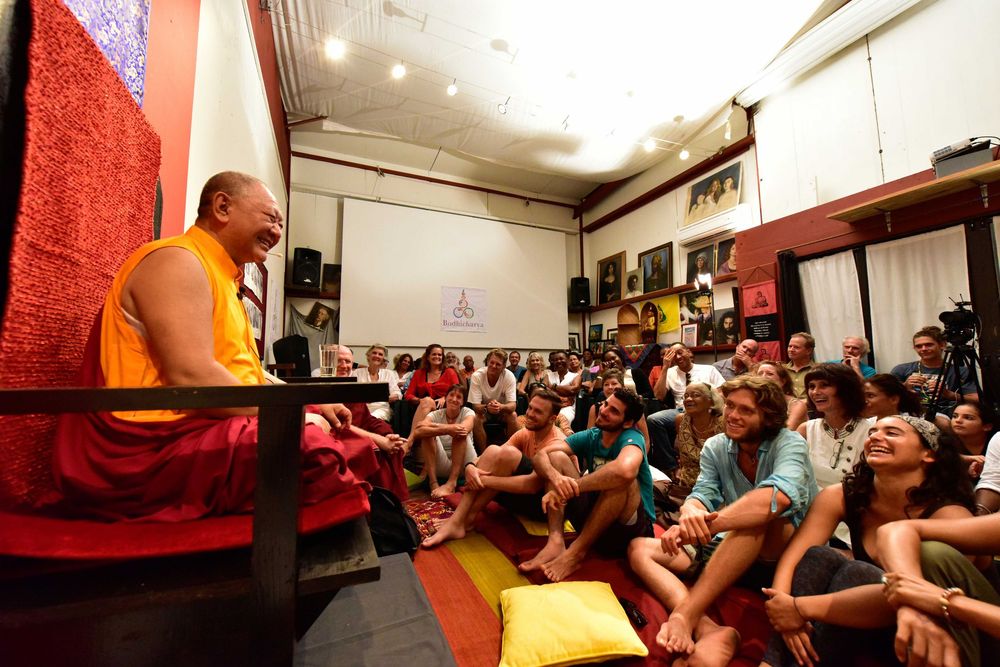 Buddhist monk in orange robes teaching a diverse group of seated students in a meditation hall.