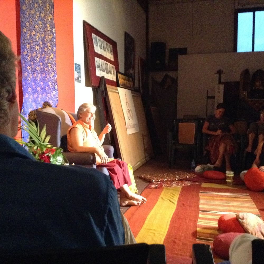 Buddhist monk in red robes reads to a seated audience in a warmly lit meditation hall with colorful decor.
