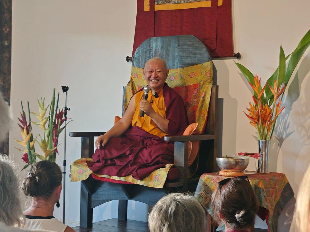 Buddhist monk in maroon robes smiles while speaking into microphone, seated on ornate chair before audience.