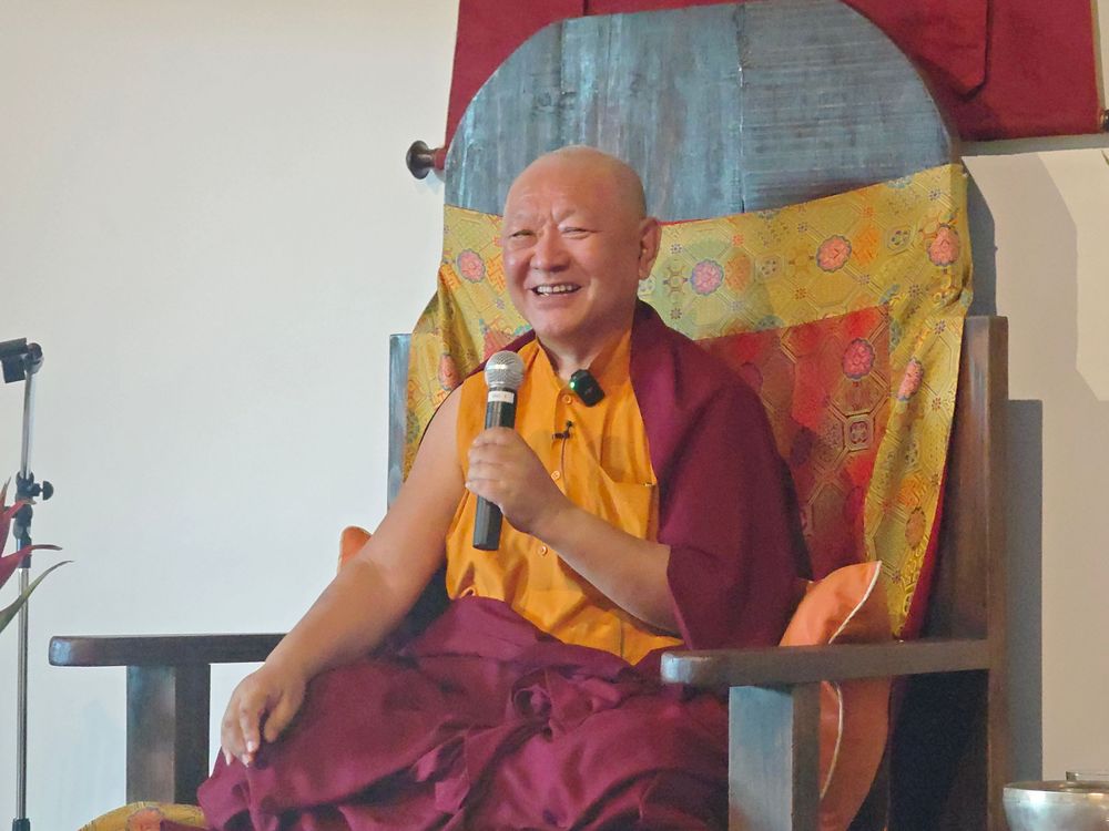 Smiling Buddhist monk in maroon and gold robes holds microphone while seated in ornate wooden chair during teaching.