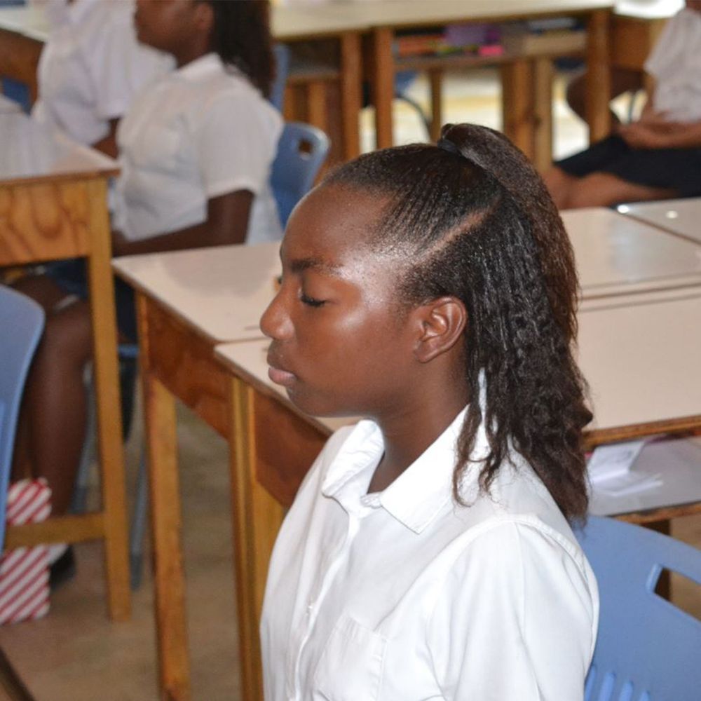 Student in white uniform sits at wooden desk with eyes closed in a classroom setting.