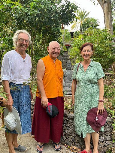 Three people smiling outdoors in a garden, including a Buddhist monk in orange robes, posing near a stone Buddha statue.