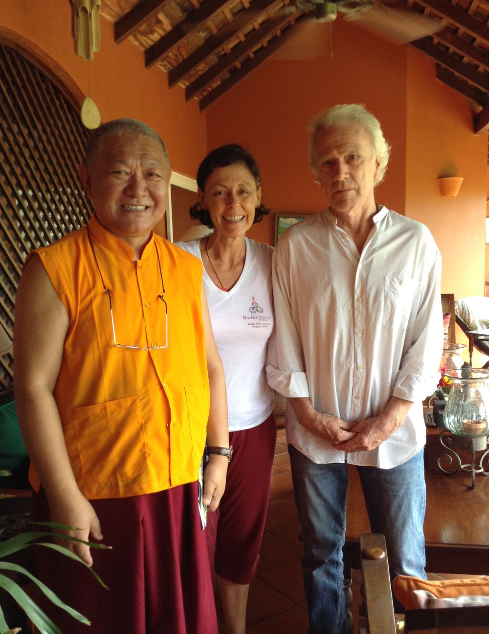 Buddhist monk in orange robes poses with two visitors inside a warm orange-walled building with wooden ceiling.