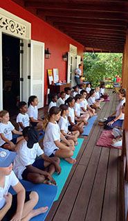 Children in white shirts practice yoga meditation on colorful mats on a red building's porch.