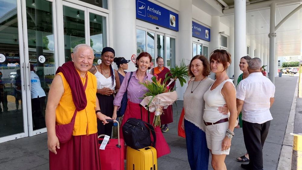 Buddhist monk in golden robes greets women with flowers outside an airport terminal, with travelers and luggage nearby.