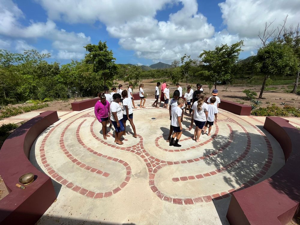 Students explore a decorative circular labyrinth mosaic outdoors, surrounded by lush greenery under a partly cloudy sky.