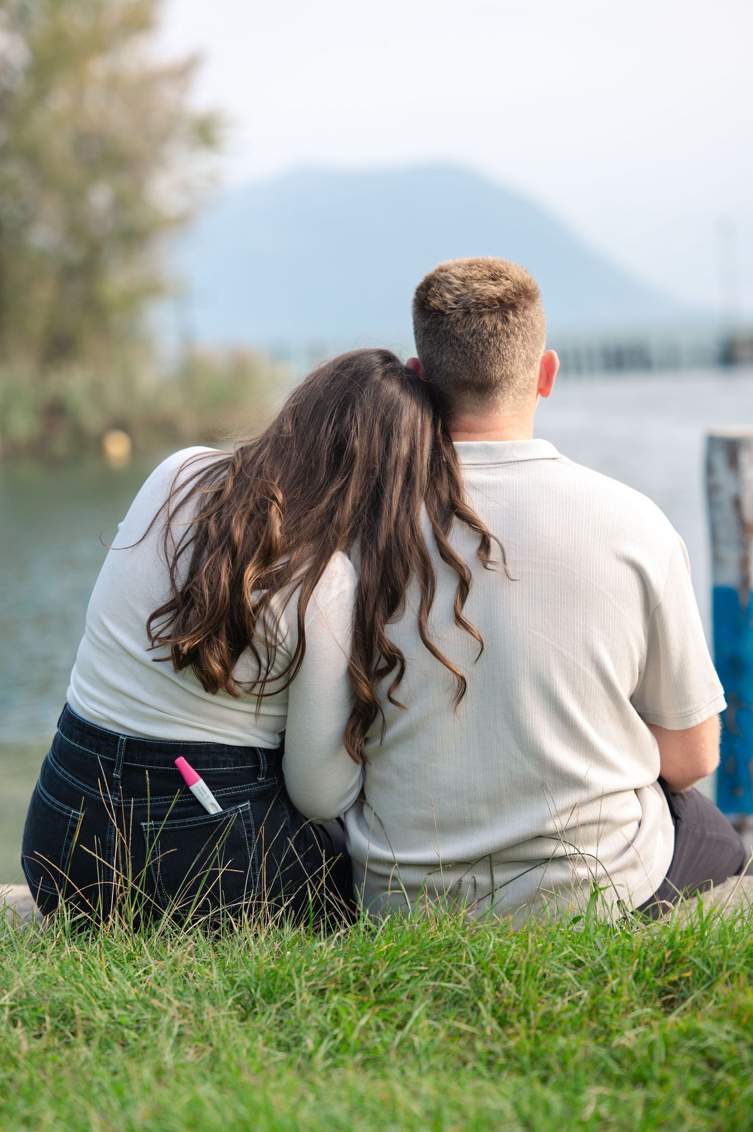 Couple sitting together by a lakeside, woman resting head on man's shoulder, mountains in background.