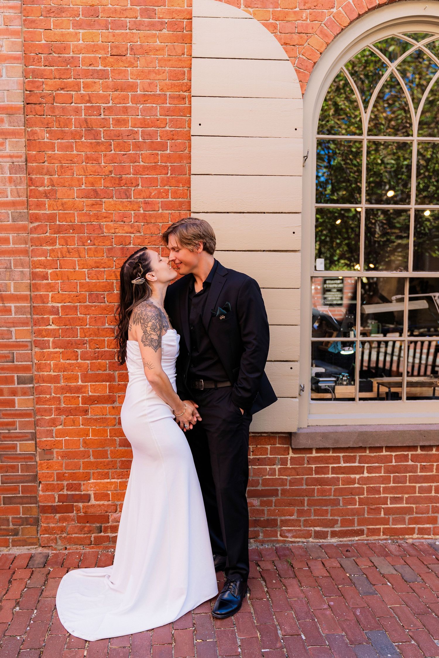 Bride reads wedding vows to groom in blue suit outdoors with city building backdrop on sunny day.